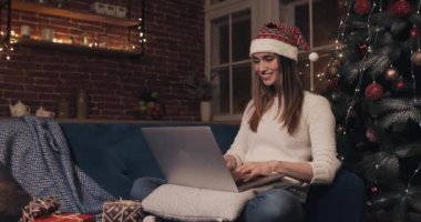 Gülümseyen Güzel Sarışın Beyaz Kız Noel Baba Şapkalı Ev Arkaplanı 'nda Noel Baba Ağacı' nın yanında oturmuş Laptop Sohbeti Yazı Tutanağı Tatil ve Yeni Yıl Tarafı Görünümü kullanıyor..