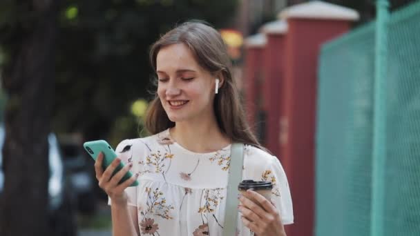 Usmívající se hezká hnědá dívka s bílými květinovými šaty a taška s sluchátky drží šálek kávy pomocí svého smartphonu. Žena poslouchající hudbu vypadá šťastně kráčející po ulici. — Stock video