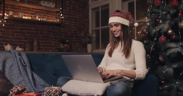 Sonriente hermosa joven rubia caucásica chica usando sombrero de santas sentado cerca del árbol de Navidad en el fondo del hogar usando su computadora portátil chateando mensajes de texto escribiendo concepto de días festivos y vista lateral de año nuevo . — Vídeo de stock