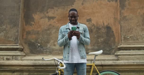 Beau Afro-Américain dans les écouteurs portant Jean Jacket en utilisant son Smartphone Moderne. Il se tient devant le vélo moderne et le mur du vieux bâtiment à l'arrière-plan Fermer . — Video