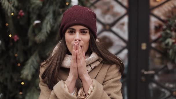 冬の帽子をかぶっている若い魅力的なブルネットの女の子の肖像カメラを見て、笑顔、温暖化、そして彼女の手をこすりますクリスマス装飾されたウィンドウショップの近くに立って閉じる。休日のコンセプト. — ストック動画