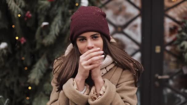 Portrait eines niedlichen brünetten kaukasischen Mädchens mit Wintermütze, das sich kalt fühlt, ihre Hände wärmt, in die Kamera blickt, vor einem weihnachtlich geschmückten Schaufenster steht. Nahaufnahme. Urlaubskonzept. — Stockvideo