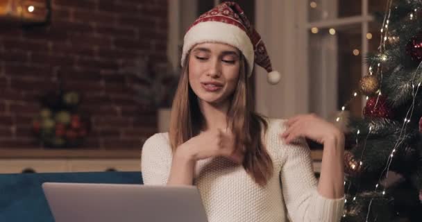 Leende kaukasiska kvinna bär Christamas hatt Sitter på soffan på Cosy Home nära julgranen gör ett videosamtal med hjälp av laptop vifta Hej visar ett träd. Begreppet semester och nyår. — Stockvideo