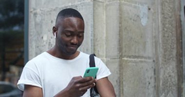 Yakışıklı Afro-Amerikan 'ın kulaklıklı, sıradan giysili ve akıllı telefonunu kullanan fotoğrafını çek. Adam Müzik Dinliyor, Heyecanlı Bakıyor Sokakta Dururken.