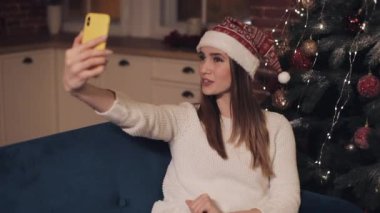 Mutlu Kafkasyalı Genç Güzel Kız Noel şapkası takıyor Noel ağacının yanındaki koltukta oturuyor Selfie yapıyor ya da görüntülü konuşma ve poz veriyor. Tatil ve Yeni Yıl kavramı.