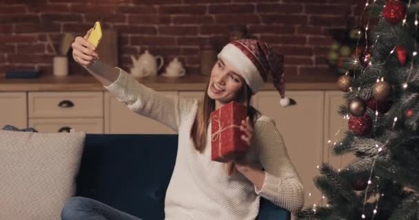 Happy Caucasian Young Pretty Girl Wearing Christamas Hat Sitting on the Sofa Holding and Shaking Present Box near Christmas Tree Making a Video Call or Video Message. Concepto de Vacaciones y Año Nuevo . — Vídeos de Stock
