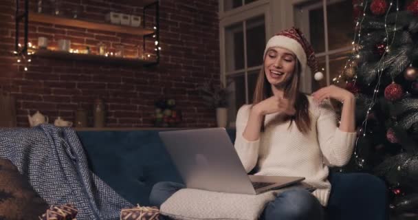 Happy Caucasian Girl Wearing Christamas Hat Sitting on the Sofa at Cosy Home Background near Christmas Tree. Mujer haciendo una videollamada usando su computadora portátil hablando mostrando un árbol . — Vídeos de Stock