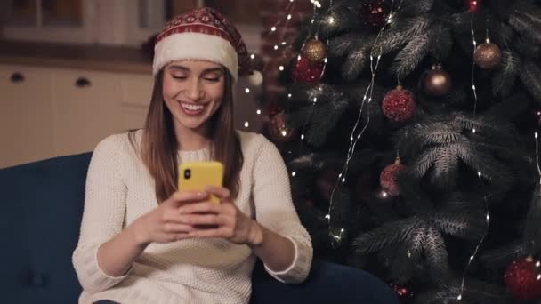 Щаслива кавказька молода дівчина, яка носить Крістамас - капелюх, сидячи на софі біля Різдвяного дерева, робить сельфі або відеодзвінок, що показує, як виглядатиме, як око зворушує своєю рукою. — стокове відео