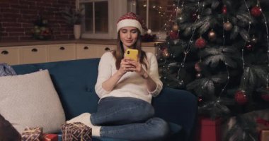 Gülümseyen Kafkas Sarışın Kız Noel Baba Şapkalı Ev Arkaplanı 'nda bir koltuğa oturmuş Smartphone tutuyor Mesaj gönderiyor ve mesajları okuyor. Tatil ve Yeni Yıl kavramı.