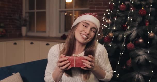 Sonriente chica bonita joven con sombrero de Santas sentado cerca del árbol de Navidad saludando Hola sosteniendo y dando un saludo de caja de regalo roja riendo soplando un beso haciendo una videollamada o mensaje de video — Vídeo de stock