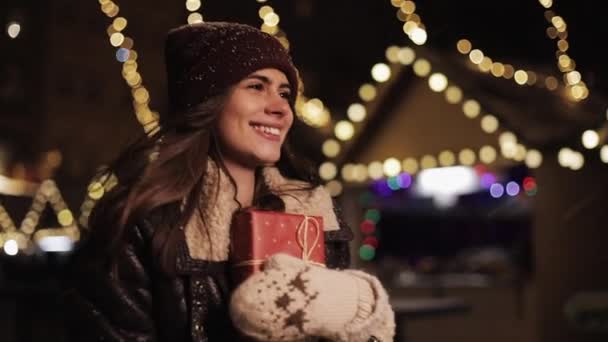 Junges süßes lächelndes Mädchen mit Wintermütze, Geschenkbox haltend, glücklich und aufgeregt aussehend, im fallenden Schnee im Hintergrund der Weihnachtsmarktbeleuchtung wandelnd. Urlaubskonzept. — Stockvideo