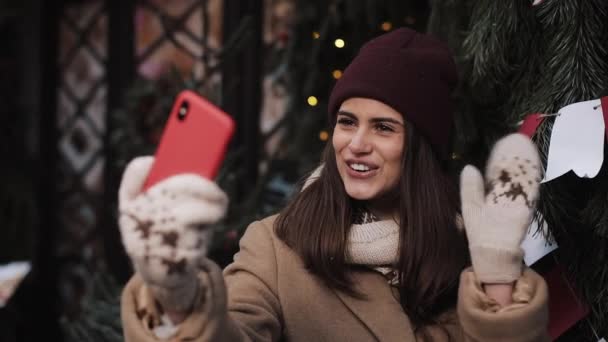 겨울 옷을 입고 포즈를 취하고 손을 흔들고 키스를 하고 크리스마스 배경으로 야외에 서 있는 아름다운 소녀가 셀카나 비디오 메시지를 만들고 있다. 겨울 휴가 개념 — 비디오