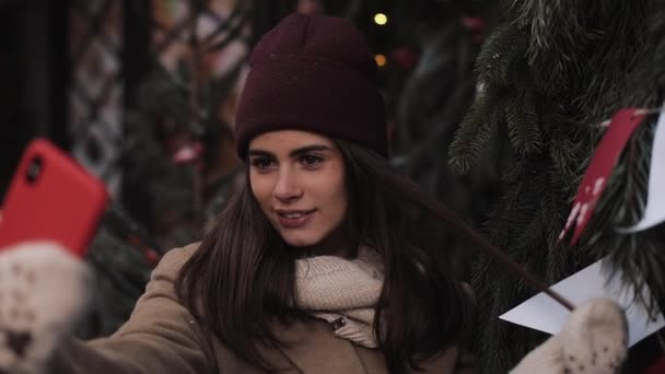 Close Up of Young Beautiful Girl Wearing Hat, Scarf, Mittens, Posing and Smiling, Making Selfie, Standing Outdoor at Christmas Decorated Background. Зима. Концепция праздника и технологии . — стоковое видео