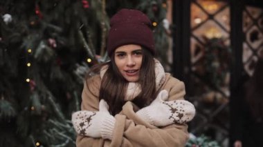 Kış giysileri giyen, kameraya bakan, çapraz ellerle dikilen, Noel süslemeli arka planda kendini ısıtan ve ovuşturan genç ve çekici kızın portresi..