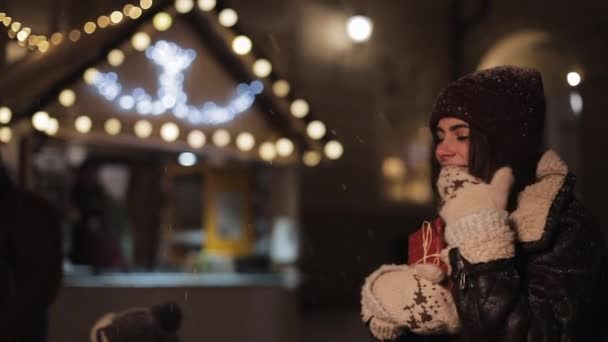 Bella ragazza sorridente che indossa il cappello invernale che tiene la scatola regalo, sembra felice ed eccitata, camminando nella neve cadente allo sfondo delle luci della fiera di Natale. Concetto Vacanze. Vista laterale destra . — Video Stock