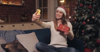 Genç Çekici Kafkas Sarışın Kız Noel Baba Şapkalı Ev Arkaplanında Bir Koltukta Oturarak Mevcut Mesaj Gönderme ve Okuma Mesajlarıyla Selfie Çekiyor.