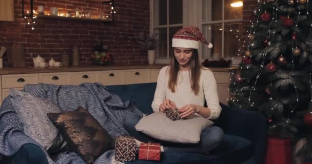 Joven chica bonita feliz con sombrero de santas sentado en el sofá cerca del árbol de Navidad Finalizar regalos de Navidad vendaje cinta y atado en un arco en el fondo de la casa — Vídeo de stock