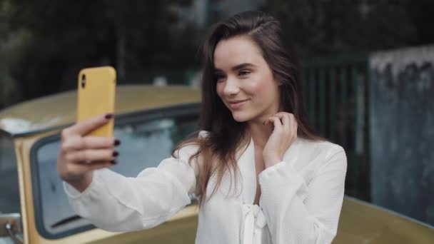 Jonge vrouw die selfie maakt op haar smartphone. Meisje poseren glimlachen en blazen van een kus overdag buiten staande leunen op Retro Golden Car. — Stockvideo