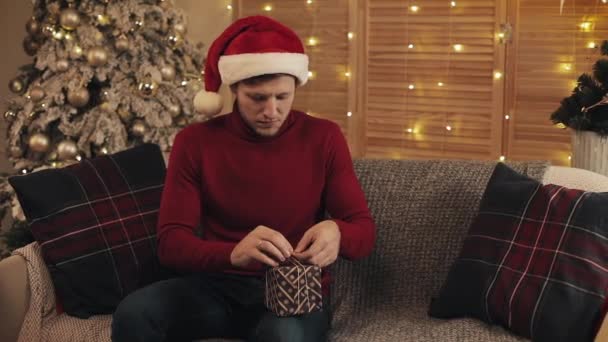 Ein junger kaukasischer Mann mit Weihnachtsmütze sitzt auf einem Sofa in der Nähe des Weihnachtsbaums im Wohnzimmer und bindet eine Schleife an eine Geschenkschachtel, blickt in die Kamera und lächelt. Konzept von Feiertagen und Neujahr. — Stockvideo