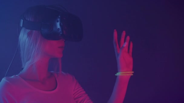 Acercamiento Retrato de una chica impresionada usando gafas de realidad virtual mirando su mano, diciendo WoW de pie en la habitación con fondo de colores de iluminación de neón . — Vídeos de Stock