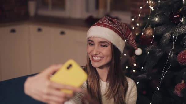 Gelukkig jong aantrekkelijk meisje dragen Christamas hoed zitten op de bank in de buurt van kerstboom maken selfie poseren zwaaien Hi Showing Tongue. Begrip vakantie en nieuwjaar. — Stockvideo