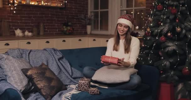 Lächelnde junge hübsche kaukasische Frauen mit Weihnachtsmütze sitzen auf einem Sofa in der Nähe des Weihnachtsbaums vor gemütlichem weihnachtlichen Hintergrund und halten eine rote Geschenkschachtel freudig in die Höhe. Konzept von Feiertagen und Neujahr. — Stockvideo