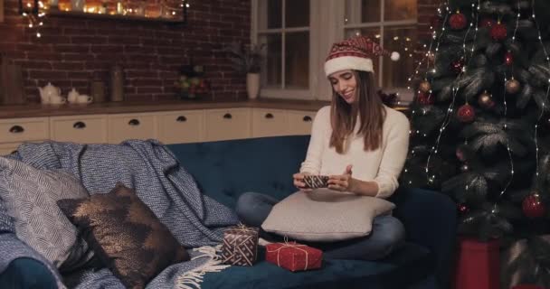 Leende ung söt kaukasiska kvinnor bär tomte hatt sitter på en soffa nära julgranen på mysig jul bakgrund håller små närvarande låda Rejoing Sätt den till två andra — Stockvideo