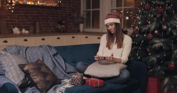 Smiling Young Pretty Caucasian Women Wearing Santas Hat Sitting on a Sofa near the Christmas Tree at Cosy Christmas Background Holding Small Present Box Rejoing Put it to two others — Stock Video