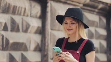 Burnunda piercing olan genç sarışın kız. Siyah şapkası var. Akıllı telefonunu kullanıyor. Eski şehrin arka planına gülümsüyor. .