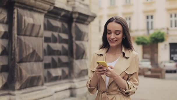 Pretty Young Girl usando una trinchera beige usando su teléfono inteligente y sonriendo disfrutando de su paseo por el fondo de la ciudad vieja . — Vídeo de stock