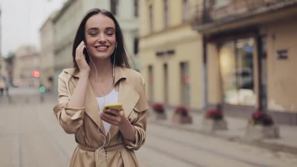 Hübsches junges Mädchen mit Kopfhörern hört gerne Musik, hält ihr Smartphone in der Hand und lächelt vor dem Hintergrund der Stadt. — Stockvideo