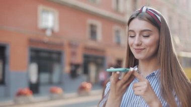 Üzerinde kahverengi saç bandı ve şeritli tişörtü olan ve akıllı telefonunu kullanan güzel beyaz bir kız. Kadın Sesli Mesaj Gönderiyor ve Kaydırıyor Şehir Caddesinde Duruyor.