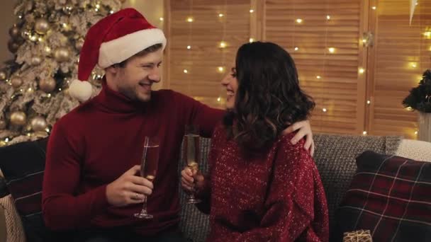 Feliz joven pareja sentada en el sofá cerca del árbol de Navidad en el acogedor hogar Antecedentes Triturar copas de champán Hablar sonriendo. Concepto de vacaciones y año nuevo de cerca . — Vídeo de stock