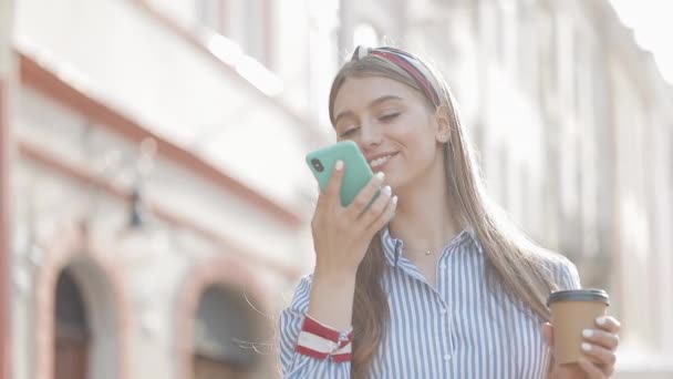 Giovane bella ragazza caucasica con capelli castani e fascia su di esso che indossa camicia a righe utilizzando il suo smartphone. Donna che invia messaggi vocali e scorre in piedi sulla City Street . — Video Stock