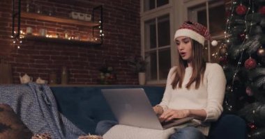 Noel Baba Şapkası Giyen Çekici Genç Kadın, Ev Arkaplanı 'nda Noel Ağacı' nın yanında oturuyor Dizüstü bilgisayarındaki Daktilo ile Harika ve Düşünceli Görünüyor..
