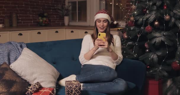 Attarctive Souriant Caucasien Blond Fille Portant Santas Chapeau Regarder heureux assis sur un canapé à la maison de Noël Arrière-plan Utilisation de son Smartphone SMS Envoi et lecture de messages disant Wow — Video