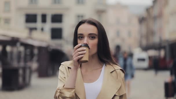 Porträt eines attraktiven jungen Mädchens, das Kaffee trinkt, stehend in die Kamera blickt und auf den Hintergrund der alten Stadt zoomt. — Stockvideo