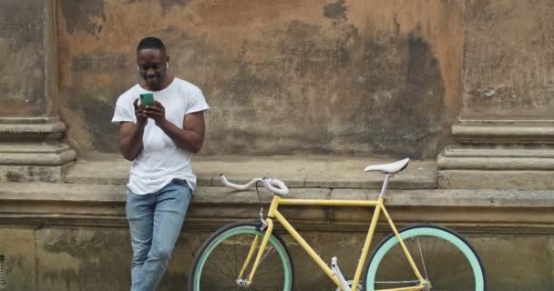 Glücklich lächelnder afroamerikanischer Mann mit Kopfhörern, der sein Smartphone benutzt, um Nachrichten in soziale Netzwerke zu tippen. männlicher Sportler steht neben stylischem modernem Fahrrad an der alten Hauswand. — Stockvideo