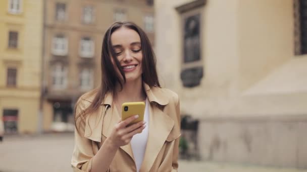 Hübsche junge Frau im beigen Trenchcoat mit ihrem Smartphone und lächelnd, während sie vor Altstadtkulisse ihre Haare fegt. Technologie und Kommunikationskonzept. — Stockvideo