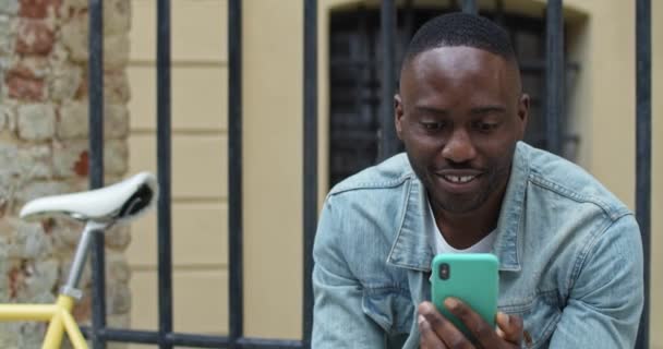 Close Up of Smiling African American Man Tendo uma chamada de vídeo através de seu Smartphone. Male Tourist Waving, sorrindo enquanto sentado perto de bicicleta elegante com Old Metal Fance no fundo. Vista frontal . — Vídeo de Stock