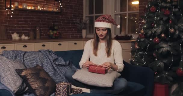 Νεαρή χαρούμενη χαμογελαστή γυναίκα με χριστουγεννιάτικο καπέλο που κάθεται στον καναπέ κοντά στο χριστουγεννιάτικο δέντρο προετοιμάζοντας δώρα και κοιτάζοντας την κάμερα στο σπίτι φόντο. Έννοια των διακοπών και το νέο έτος. — Αρχείο Βίντεο