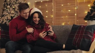 Genç ve güzel bir adam güzel karısına sarılıyor Noel ağacının yakınındaki koltukta oturmuş Smartphone Screen 'e bakan parlak ışıklarla gülümsüyor..