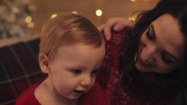 Arkaplanda Parıldayan Işıklarla Oturan ve Annelerin Dizlerine Atlayan Sevimli Küçük Gülümseyen Çocuk 'un Yakın Çekimi. Aile Tatili ve Yeni Yıl Konsepti.