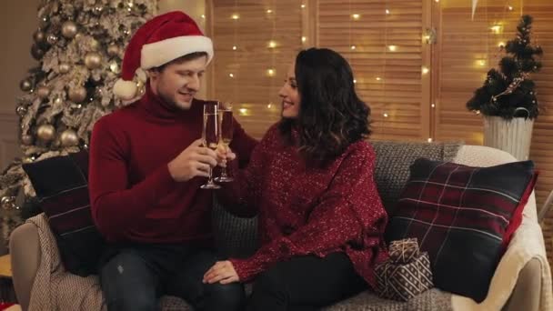 Schönes junges Paar sitzt auf dem Sofa in der Nähe des Weihnachtsbaums zu Hause Hintergrund küssen und heben Gläser Champagner. — Stockvideo