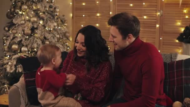 Ευτυχισμένοι γονείς που κάθονται στον καναπέ κοντά στο χριστουγεννιάτικο δέντρο στο Cosy Home Background Κρατώντας το Ενθουσιασμένο Μωρό Γιος Παίζοντας μαζί του Χαμογελώντας. Έννοια των οικογενειακών διακοπών και το νέο έτος. — Αρχείο Βίντεο