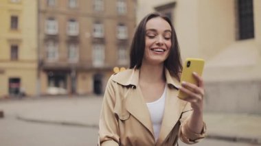 Kahverengi saçlı, bej rengi siperli, akıllı telefonuyla telefon görüşmesi yapan, Eski Şehir Arkaplanına gülümseyen güzel bir kız..