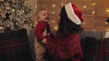 Genç, güzel anne Noel Baba şapkası takıyor, tatlı bebeğiyle koltukta oturuyor ve Noel ağacının yanındaki tatil evinde onu öpüyor.