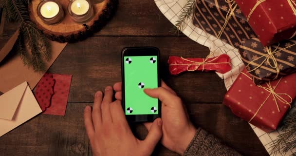 Von oben: Männliche Hände wischen den vertikalen grünen Bildschirm eines schwarzen Smartphones auf einem Holztisch hoch, der mit Geschenken und Kerzen dekoriert ist. Chroma-Schlüssel. Weihnachtsferien-Konzept. — Stockvideo