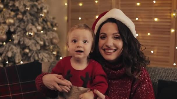 Kersttijd. Gelukkig jonge moeder en zoon kijken naar je poseren in de camera zittend op een bank in de woonkamer thuis. Kerstmis en Nieuwjaar concept. — Stockvideo