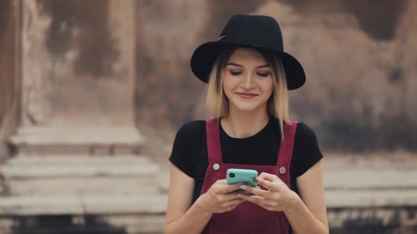 Молодая Прити улыбающаяся блондинка с пирсингом в носу в черной шляпе с помощью смартфона печатает и смеется, гуляя по городской улице  . — стоковое видео